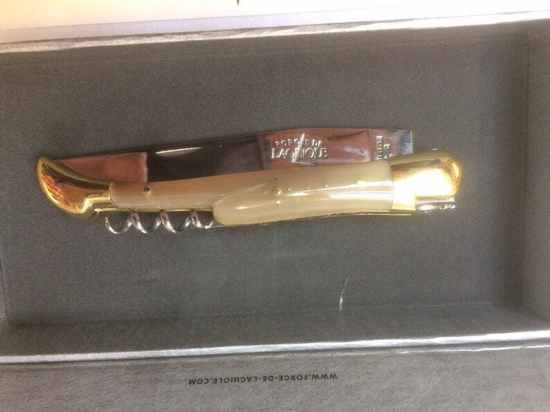May include: A gold and ivory-colored pocket knife with a corkscrew and the words "Forge de Laguiole" on the blade. The knife is in a box with the website address "www.forge-de-laguiole.com".