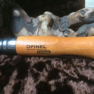 May include: A brown wooden handled Opinel knife with a silver blade. The knife has the text "Opinel Savoie - France Carbone" engraved on the handle.