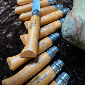 May include: A close-up of a group of wooden-handled pocket knives with the brand name "Opinel" printed on the handles. The knives are arranged in a row, with the blades folded closed. The knives are made in France and have a carbon steel blade.