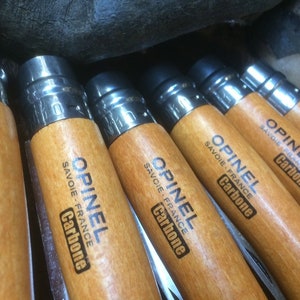 May include: Close-up of five Opinel pocket knives with wooden handles. The knives have the text "Opinel Savoie France Carbone" printed on the handles.