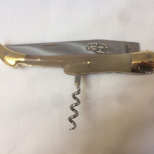 May include: A brown handled pocket knife with a corkscrew, bottle opener, and a blade. The knife has a gold colored handle and the words "Forge de Laguiole" are engraved on the blade.