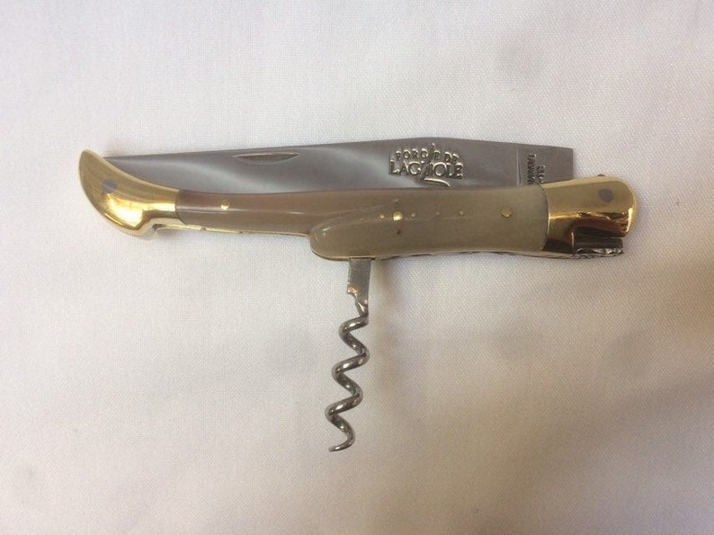 May include: A brown handled pocket knife with a corkscrew, bottle opener, and a blade. The knife has a gold colored handle and the words "Forge de Laguiole" are engraved on the blade.
