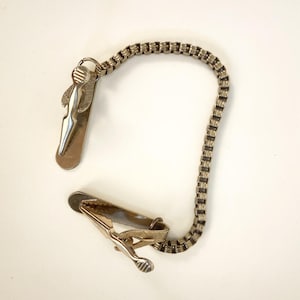 Könnte beinhalten: Eine silberfarbene Krawattenklammer mit Kette. Die Klammer hat eine rechteckige Form mit einem dekorativen Design. Die Kette besteht aus kleinen, rechteckigen Gliedern. Die Krawattenklammer dient dazu, eine Krawatte an Ort und Stelle zu halten.