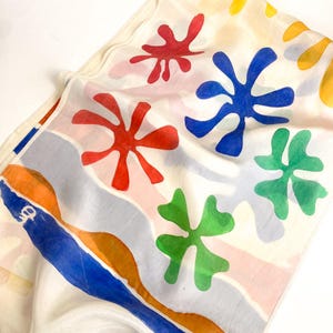 May include: A white silk scarf featuring a colorful abstract pattern of red, blue, and green organic flower-like shapes. The design includes wavy bands of orange, blue, and light gray along one edge, with a small white logo printed on the blue stripe.
