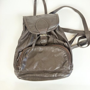 May include: A dark brown leather backpack featuring a top flap with a buckle closure, a drawstring opening, and a front zippered pocket. The bag includes adjustable shoulder straps and a top handle, showcasing a soft, textured finish.