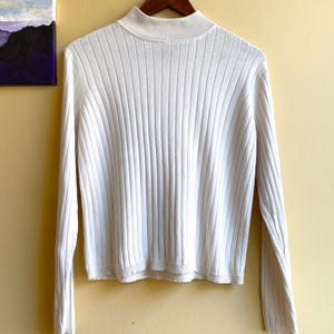 Pode incluir: Um suéter branco de manga comprida com gola alta e um padrão de malha canelada vertical. A peça tem uma bainha reta e mangas ajustadas, confeccionada em tecido de malha de cor sólida com uma textura consistente em toda a sua extensão.