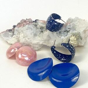 Könnte beinhalten: Eine Schmuckkollektion bestehend aus zwei dunkelblauen Creolen mit weißem Sprenkelmuster, zwei runden, blassrosa Cabochons mit Perlmutteffekt und zwei einfarbigen, leuchtend blauen runden Scheiben, die neben einem Stück Rohkristall liegen.