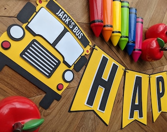 May include: A yellow school bus cutout with the words "Jack's Bus" on the side. A yellow banner with black letters spelling "Happy" is hanging below the bus. There are red apples and colorful crayons on a wooden surface.