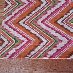 May include: A colorful table runner with a chevron pattern in shades of orange, pink, green, and brown. The runner is made of a woven fabric with a slightly textured surface.