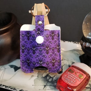 May include: Purple fabric hand sanitizer holder with a Haunted Mansion pattern. The holder has a snap closure and a white button. A small bottle of hand sanitizer is in the foreground. The bottle is red and has the text "Champagne Apple & Honey" on the label.