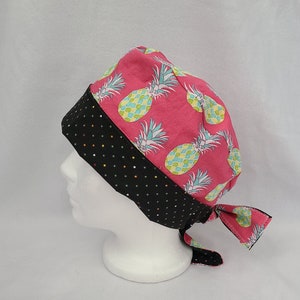 May include: A pink scrub cap with a repeating pattern of green and blue pineapples. The cap has a black polka dot band and ties. The cap is designed to be worn on the head.