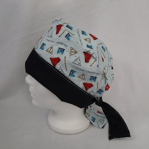 May include: A light blue bandana-style hat featuring a pattern of red, blue, and gold symbols and text. The hat has a black band and trim, and is tied at the back. The hat is displayed on a white mannequin head.