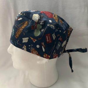 Può includere: Una cuffia chirurgica blu scuro con un motivo ripetuto di illustrazioni a tema golf, tra cui sacche, mazze, palline, scarpe e cartelli rossi con la scritta CLUBHOUSE. La cuffia è dotata di una chiusura a laccetti sul retro.