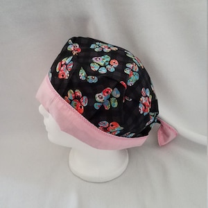 Op de afbeelding: Een zwarte en roze chirurgische scrubcap met een bloemenpatroon. De cap heeft een zwart geruite stof met kleurrijke bloemmotieven en een roze rand. De cap is aan de achterkant vastgebonden.