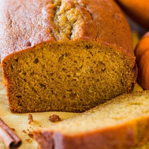 May include: A loaf of pumpkin bread sliced in half, with a slice of bread in front of it. The bread is a light brown color and has a slightly crumbly texture. There are some cinnamon sticks in the background.