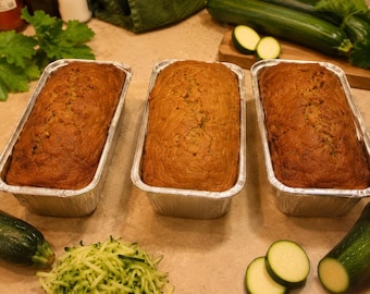 Op de afbeelding: Drie goudbruine courgettebroden gebakken in rechthoekige aluminium bakvormen. De broden liggen op een oppervlak naast hele groene courgettes, schijfjes courgette en een hoopje geraspte groene courgette.