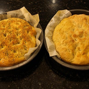 May include: Two round loaves of baked bread in dark metal pans lined with parchment paper. The left loaf is golden brown with a textured surface topped with green herbs. The right loaf is a smooth, pale golden brown with a dimpled surface.