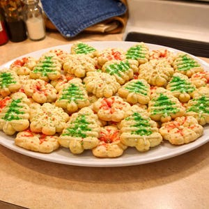 May include: A large white platter filled with assorted holiday cookies. The collection includes light-colored, textured cookies shaped like Christmas trees decorated with green icing, and round, flower-shaped cookies with red sugar sprinkles.