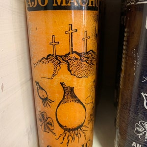 May include: Orange candle with a black label that says "AJO MACHO GARLIC". The label also has a drawing of three crosses, a garlic bulb, and three shamrocks.