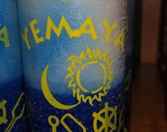 May include: A blue and white candle with a yellow design. The candle has the words "Indi's Orishas Yemaya" written on it. The design includes a crescent moon, a sun, an anchor, and a wheel.