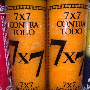 May include: Two orange candles with the text "7x7 Contra Todo" and "7x7 Against Everything" printed on them. The candles are in glass jars with a black label.