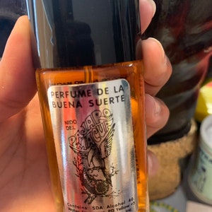 Puede incluir: Un pequeño frasco de perfume con tapa negra y etiqueta plateada. La etiqueta dice "PERFUME DE LA BUENA SUERTE" y presenta una ilustración de un pájaro. El frasco contiene un líquido ámbar claro.