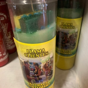 以下が含まれることがあります： 緑と青のワックス層を持つ2本の黄色いキャンドル。キャンドルには、スペイン語と英語で「Llama Clientes」と「Attract Customers」と書かれたラベルが付いています。