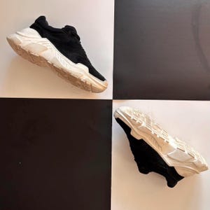May include: Black and white athletic shoe with a chunky, off-white sole. The shoe's upper is black, with a suede-like texture. The sole has a geometric design. The shoe is displayed on a black and white checkered background.
