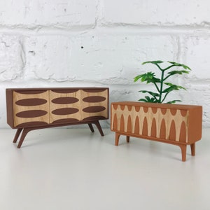 May include: Two miniature mid-century modern style cabinets. One is brown with three light-colored oval designs, and the other is orange-brown with a series of light-colored vertical designs. Both have angled legs and are set against a white brick wall.