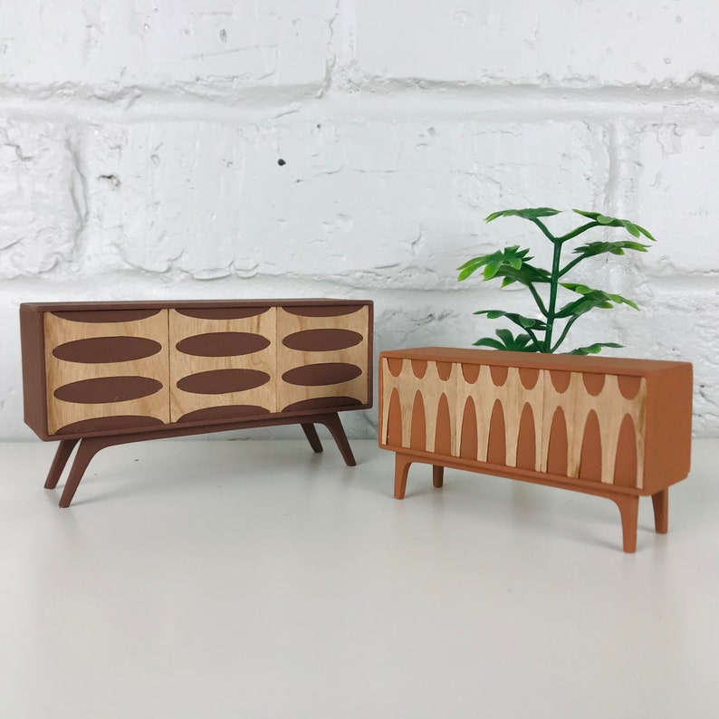 May include: Two miniature mid-century modern style cabinets. One is brown with three light-colored oval designs, and the other is orange-brown with a series of light-colored vertical designs. Both have angled legs and are set against a white brick wall.