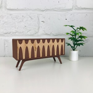 May include: A miniature brown cabinet with three doors and light wood-colored decorative panels. The cabinet has angled legs and a mid-century modern design. A small potted plant is to the right of the cabinet.