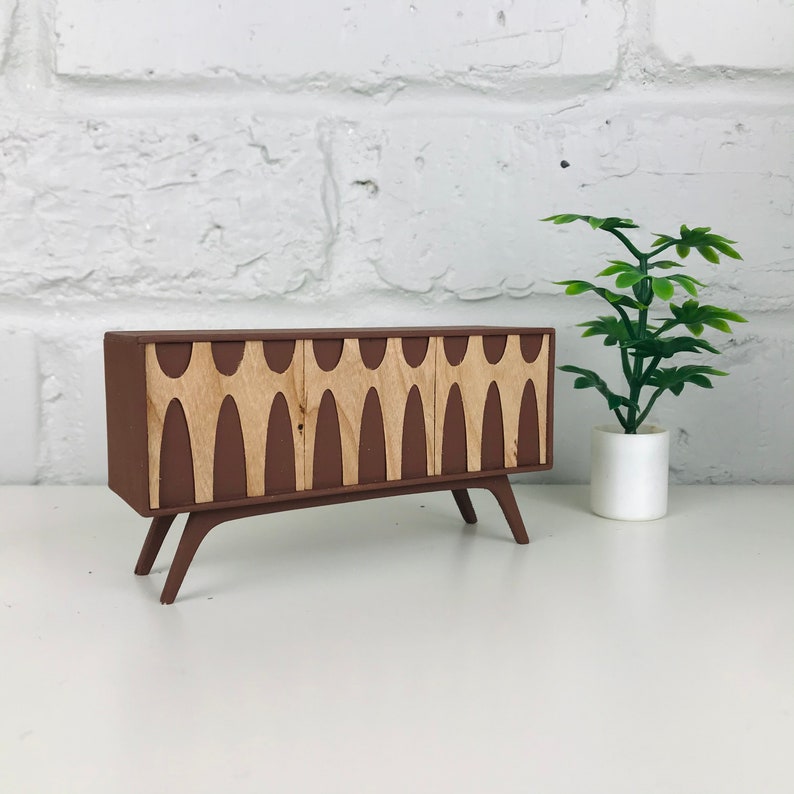 May include: A miniature brown cabinet with three doors and light wood-colored decorative panels. The cabinet has angled legs and a mid-century modern design. A small potted plant is to the right of the cabinet.