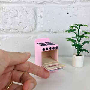 Może przedstawiać: Miniaturowy r&oacute;żowy piec z otwartymi drzwiczkami piekarnika. Piekarnik ma cztery czarne pokrętła palnik&oacute;w i cztery czarne pierścienie palnik&oacute;w. Drzwiczki piekarnika mają srebrny uchwyt.