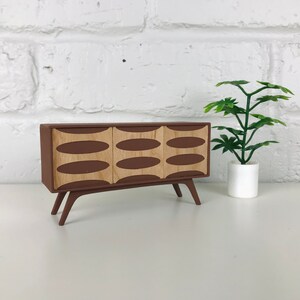May include: A miniature mid-century modern style cabinet in brown with three doors. Each door features a light wood oval design. The cabinet has angled legs and is displayed next to a small potted plant.