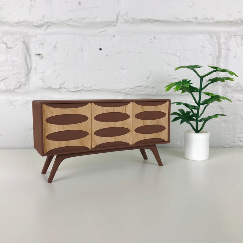May include: A miniature mid-century modern style cabinet in brown with three doors. Each door features a light wood oval design. The cabinet has angled legs and is displayed next to a small potted plant.