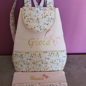 May include: Pink and white backpack with a floral pattern and the name "Gioia" embroidered in gold. The backpack comes with a matching pink and white pencil case.