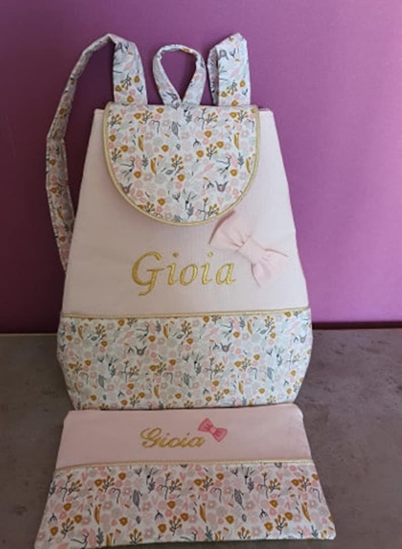 May include: Pink and white backpack with a floral pattern and the name "Gioia" embroidered in gold. The backpack comes with a matching pink and white pencil case.