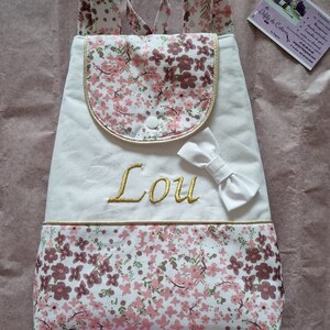 May include: White canvas backpack with a floral pattern. The backpack has a small pocket on the front with a gold trim and a white bow. The name "Lou" is embroidered in gold on the front of the backpack.