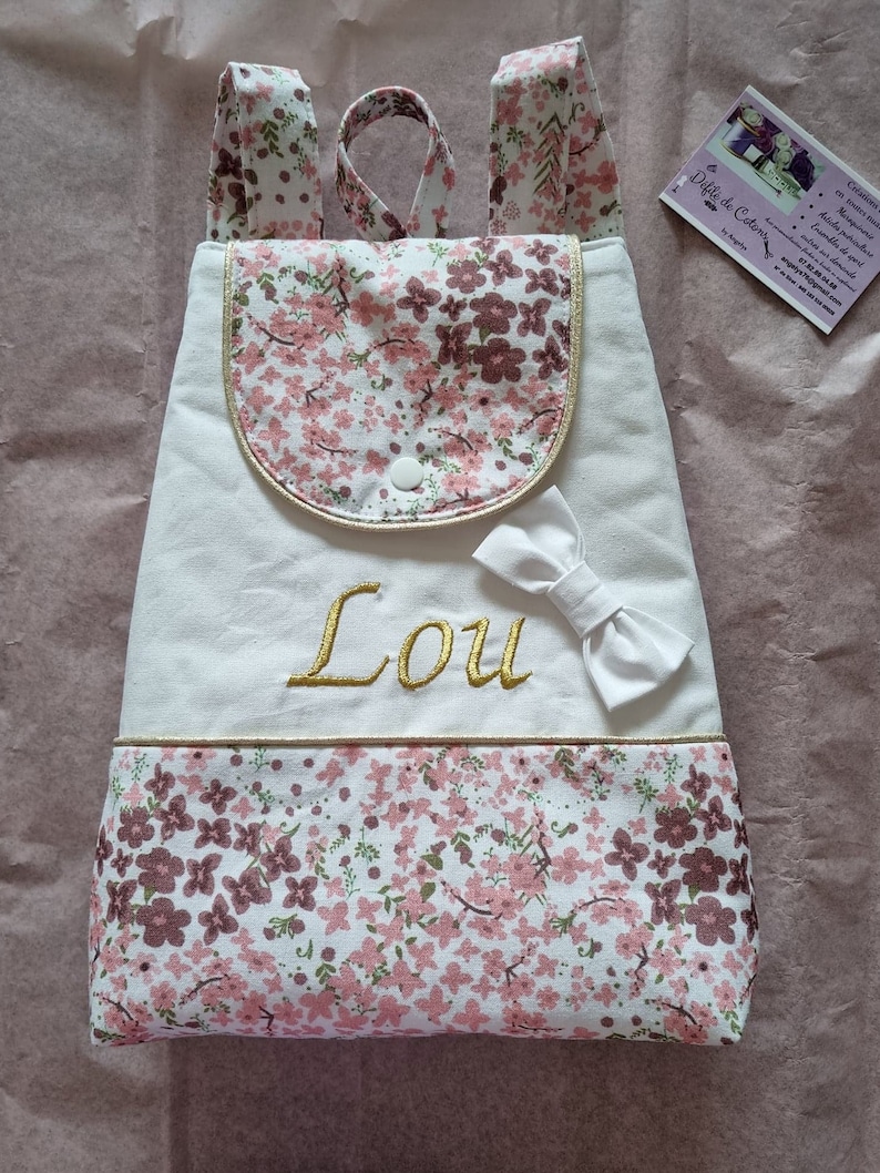May include: White canvas backpack with a floral pattern. The backpack has a small pocket on the front with a gold trim and a white bow. The name "Lou" is embroidered in gold on the front of the backpack.