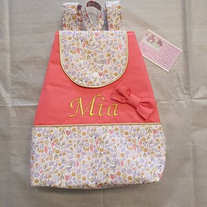 May include: A pink personalised backpack with a floral pattern and a gold trim. The backpack has a small pocket on the front and a bow on the top. The name "Mia" is embroidered on the front in gold thread.