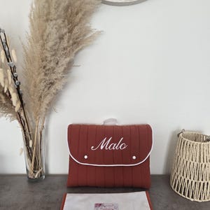 May include: A rust-colored baby changing pad and matching diaper bag with the name "Malo" embroidered in white. The bag has a white trim and a handle. A white and rust-colored changing pad is below the bag.
