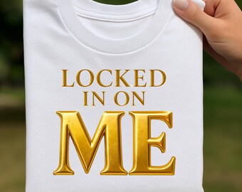 May include: White crew neck t-shirt with the gold text "LOCKED IN ON ME". The text is in a bold, serif font. The shirt is held up by a hand, with a blurred green background.