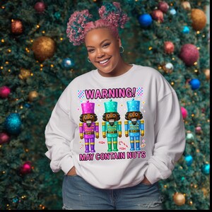 May include: White sweatshirt with a graphic of three colorful nutcracker soldiers. The text "WARNING! MAY CONTAIN NUTS" is printed above and below the nutcrackers. The background is a Christmas tree with colorful ornaments.