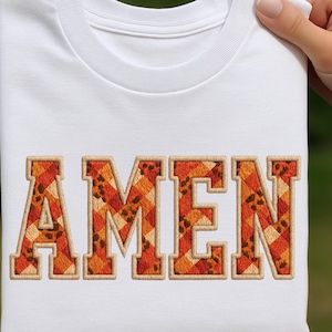 Op de afbeelding: Wit T-shirt met het woord "AMEN" geborduurd in een vetgedrukte lettertype in collegestijl. De letters hebben een patchwork-ontwerp in tinten oranje, bruin en beige, met een getextureerde, gestikte rand. Het shirt is opgevouwen.