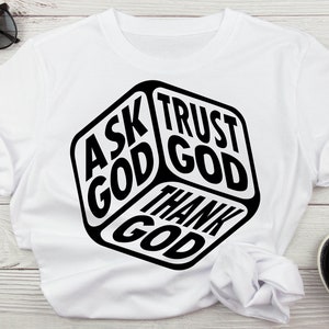 May include: White t-shirt with a black graphic of a cube with the words "Ask God", "Trust God", and "Thank God" on each side.