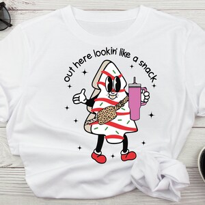 May include: White t-shirt with a cartoon gingerbread man holding a pink cup. The gingerbread man is wearing a leopard print belt and has the text "out here lookin' like a snack" above it.