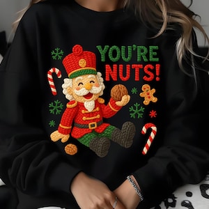 May include: Black sweatshirt featuring a colorful Christmas-themed graphic. The design includes a smiling nutcracker holding a walnut, candy canes, snowflakes, and the text "YOU'RE NUTS!" in green and red. A gingerbread man is also featured.