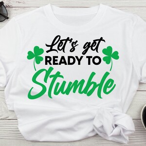 May include: White t-shirt with black text that reads "Let's get ready to stumble" with green shamrock graphics on either side of the text.