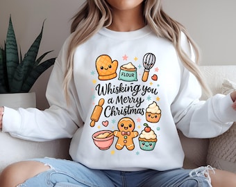May include: White sweatshirt with a colorful graphic featuring baking-themed illustrations and the text "Whisking you a Merry Christmas." The design includes a whisk, rolling pin, gingerbread man, and cupcakes.