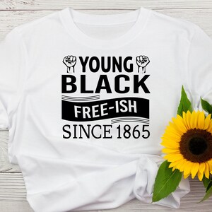 May include: White t-shirt with the text "YOUNG BLACK FREE-ISH SINCE 1865" in black. The design includes a raised fist graphic above the words "YOUNG BLACK". Yellow sunflowers are in the lower right corner.